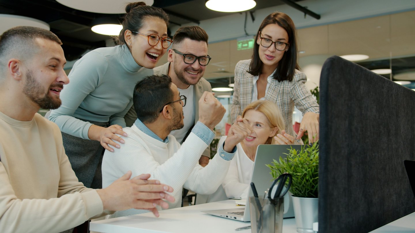 Diverse team celebrates success around a laptop in a modern office.