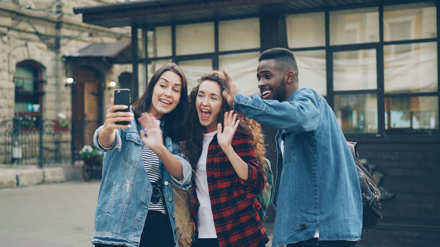 Three friends wave and smile during a video call outdoors.